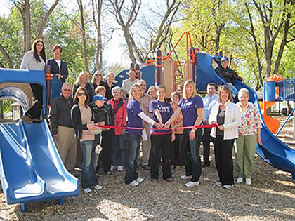 Group of people at a ribbon-cutting ceremony in front of a colorful playground structure in a park