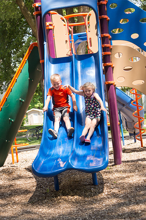 Two young children, a boy and a girl, sliding down a blue playground slide together in a vibrant park